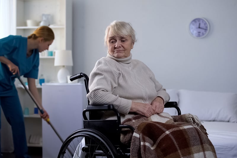 A senior woman in a wheelchair looking a little sad, alone in her room looking over her shoulder with an out-of-view support worker behind her cleaning the floor