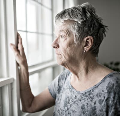 A lonely senior woman looking outside with her arm resting against the window